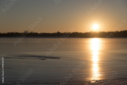 The sun sets over of frozen lake, without snow cover, so the sun reflects brightly off of it.