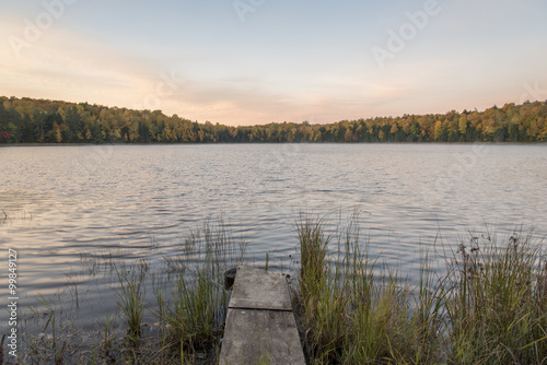 A lake with a surrounding forest in fall colors in Wisconsin.  A small wooded dock extends from the shore.
