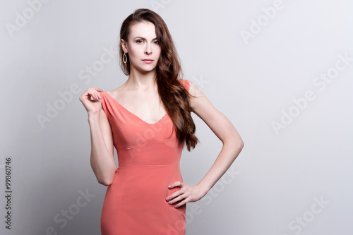 Portrait of a young attractive woman with beautiful long brown hair. She wears a coral dress and golden earrings. She touches her dress in an elegant gesture.