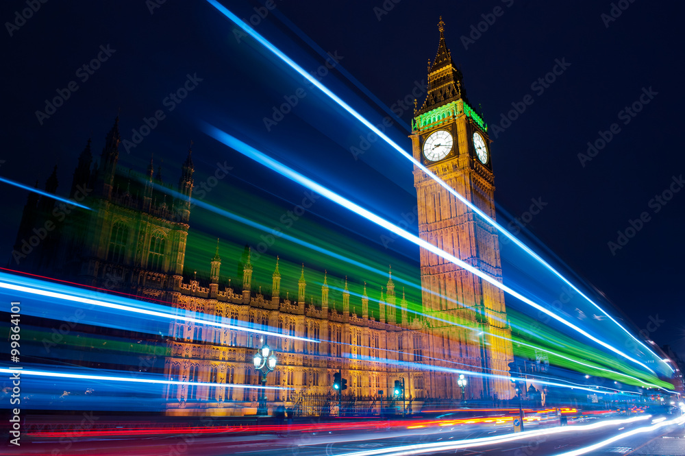 Traffic through London. Big Ben, one of the most prominent symbols of both London and England, as shown at night along with the lights of the cars and buses passing by.