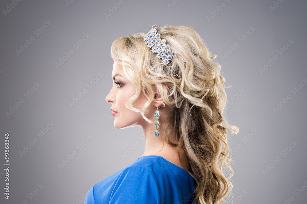Side view of beautiful, young lady wearing sapphire coronet and earrings over grey background.