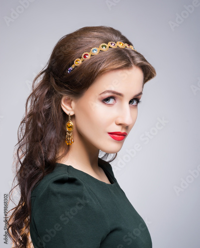 Close-up of gorgeous, young brunette with enigmatic face wearing luxury golden headband and earrings over isolated background.