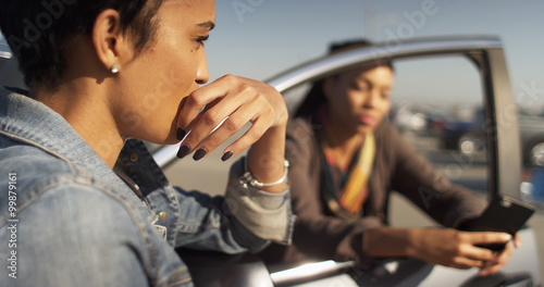 Two black women friends leaning against car talking and texting