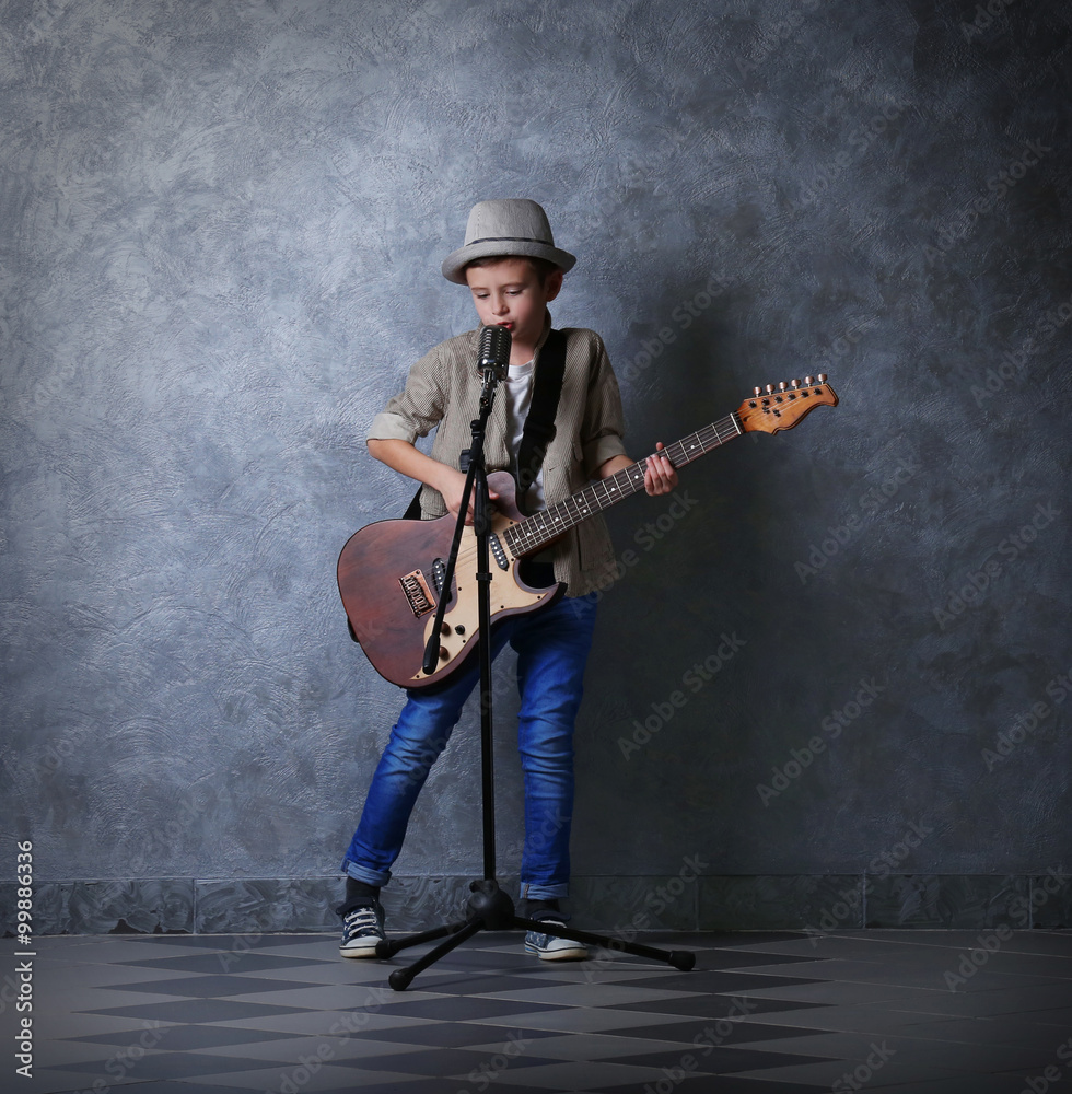 Little boy playing guitar and singing with microphone on a grey wall background Little boy playing guitar and singing with microphone on a grey wall background