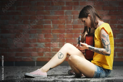 Attractive woman with tattoo playing guitar on brick background