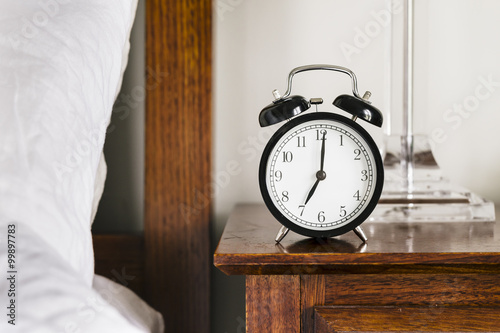 Fényképezés Black alarm clock showing 7 o'clock on bedside table