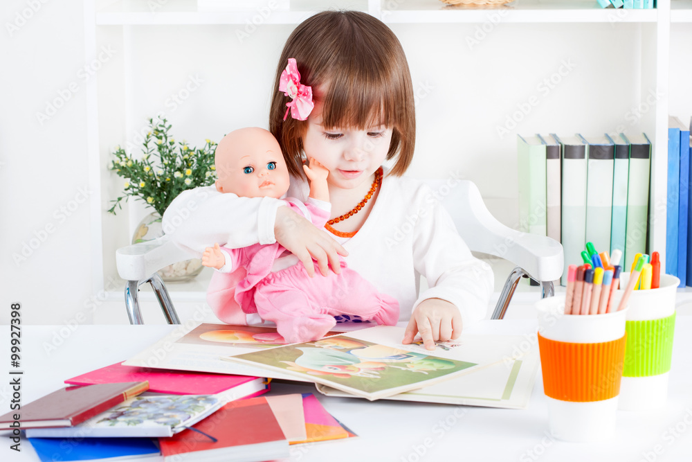 Fototapeta premium Little girl is reading a story for her baby doll while sitting by the table full of books