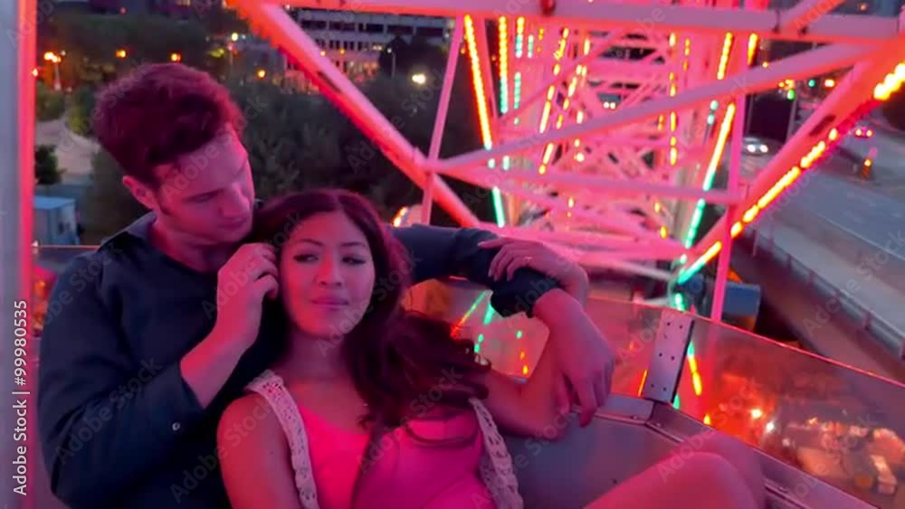 An attractive couple cuddled together on a ferris wheel at night, city buildings behind them An attractive couple cuddled together on a ferris wheel at night, city buildings behind them