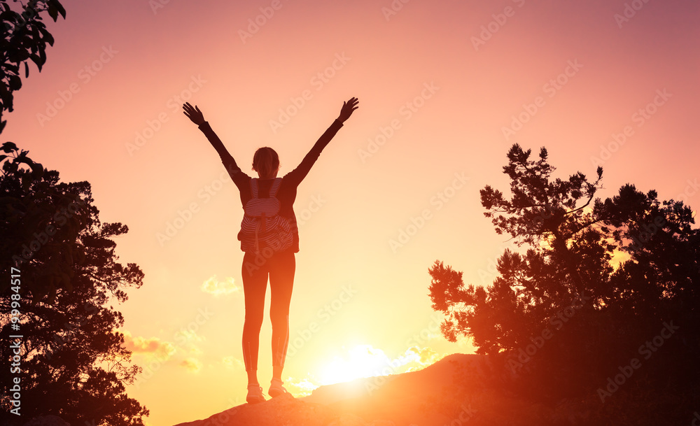 Silhouette of a happy young woman with backpack and raised arms at the sunset in mountains at summer. Nature background