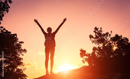 Silhouette of a happy young woman with backpack and raised arms at the sunset in mountains at summer. Nature background © den-belitsky