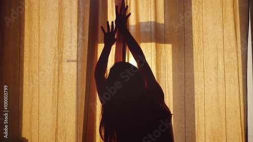 Attractive young woman is open curtains,  looking at amazing sunset above urban city standing near the window  in slowmotion