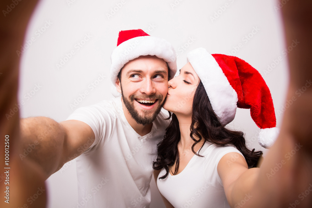 Obraz premium christmas, holidays, technology and people concept - happy couple in santa hats taking selfie picture with smartphone on white background