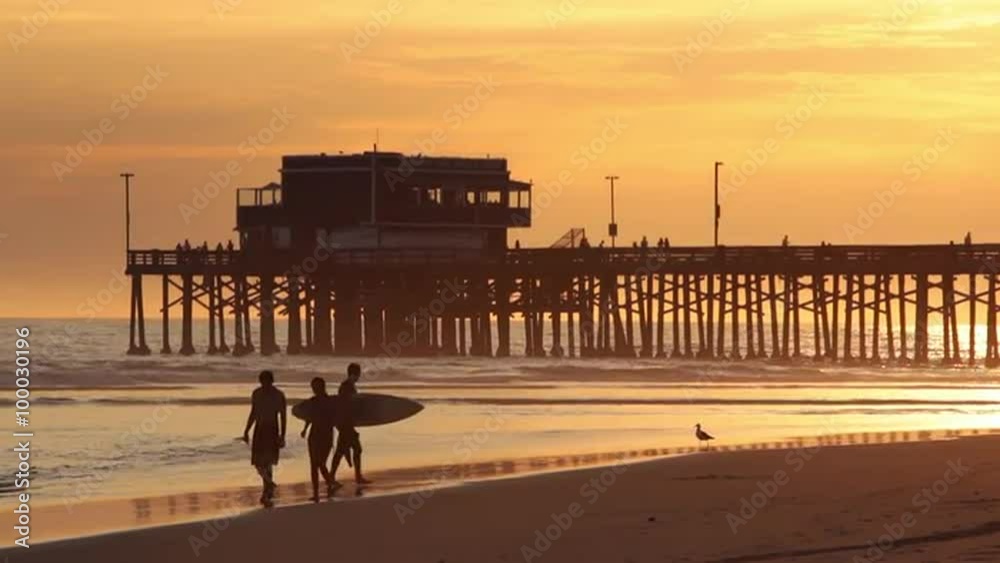 Beach sunset with silhouetted couple and surfer walking at Newport Pier, California.  