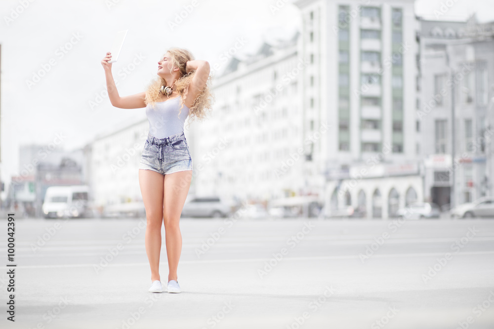 Music and selfie / Happy young woman with vintage music headphones around her neck, taking selfie with tablet pc and smiling happily against urban city background.