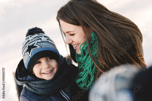 Cute young pregnant mother and her child having fun in winter park on a bright day hugging each other and smiling
