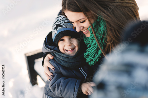 Cute young pregnant mother and her child having fun in winter park on a bright day hugging each other and smiling
