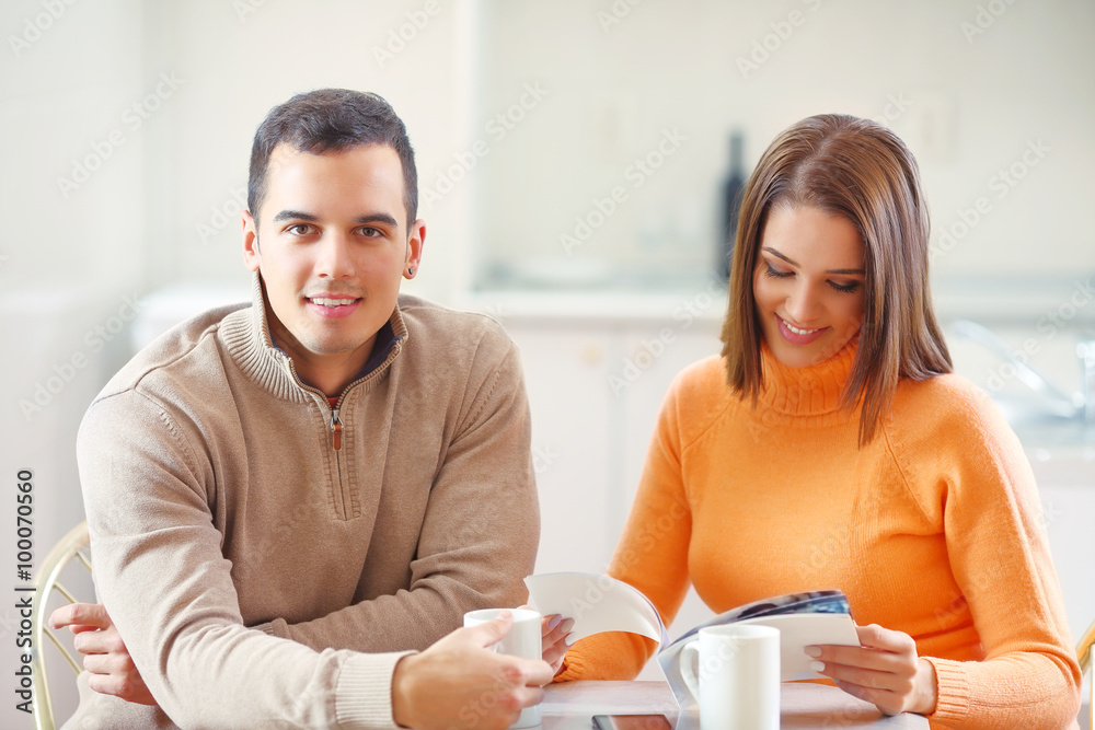 Happy young man drinking coffee at home with his girlfriend or wife who is reading a magazine Happy young man drinking coffee at home with his girlfriend or wife who is reading a magazine