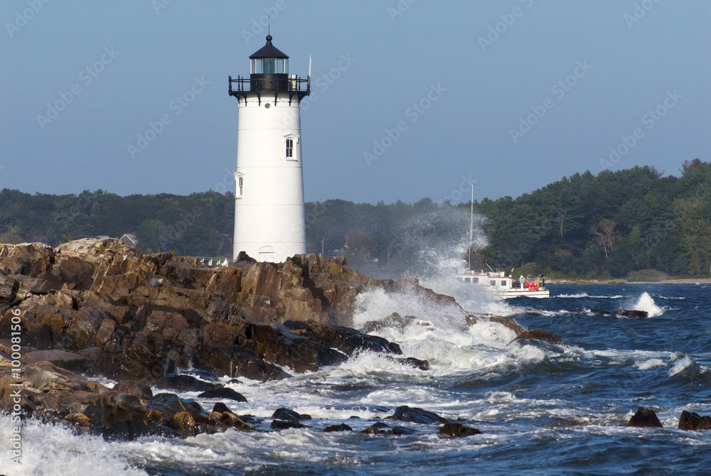 Portsmouth Harbor lighthouse guides fishing boat as waves break over rocky shore during high tide in New Hampshire. The lighthouse is also referred to as  Fort Constitution light.