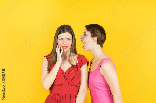 two women whispering gossip or bad news. Isolated studio yellow background female model.