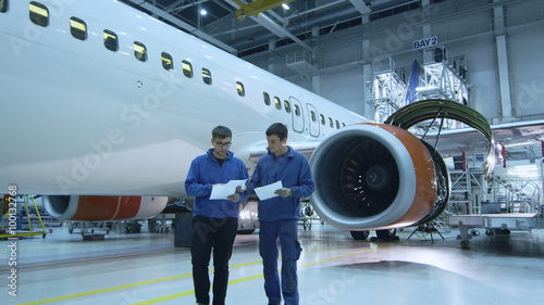 Two aircraft maintenance mechanics have a conversation while checking documents in a plane hangar with an airplane in the background. Shot on RED Cinema Camera.