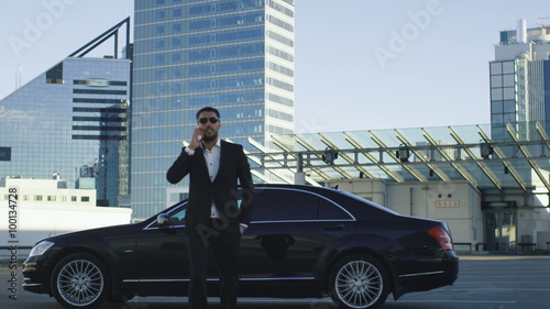 Businessman gets out of a black executive car at roof car park during day time while speaking on a phone.
