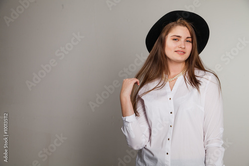 Awesome Caucasian attractive shy sexy female model with brunette hair posing on table in studio, wearing formal suit and hat, isolated on white background