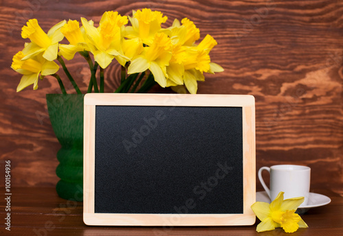 Yellow fresh daffodils in a green vase on old rustic table with chalk black bord and cup of coffee. Still life with a bouquet of narcissuses. Chalkboard menu.