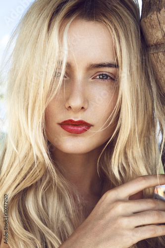 Around the palm.
Close-up portrait of beautiful young woman with curly long blonde hair, green mysterious eyes and red sexy big lips posing next to the palm and looking to you through lens.