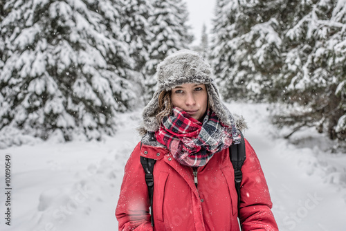 Outdoor portrait of young woman, looking at the camera. Snow covered pine trees on the background. 