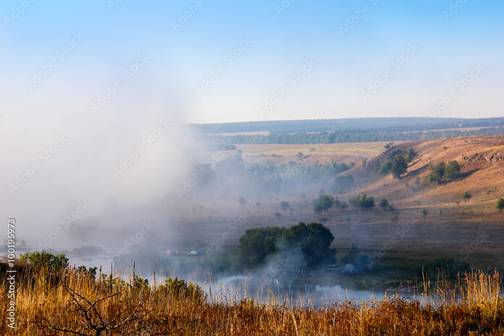 landscape of a yellow hills, trees, sky and river with dense fog