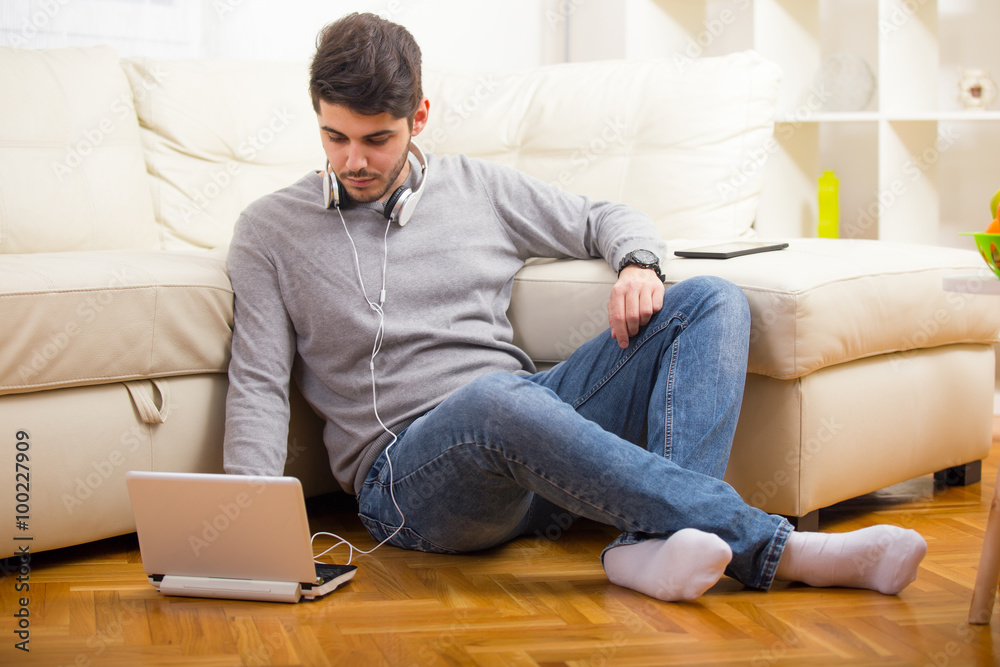 Man listening to music with earphones on laptop computer, sitting on the floor near the sofa