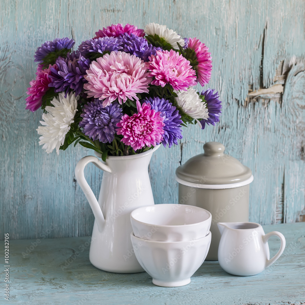 flowers asters in a white enameled pitcher and vintage crockery - ceramic bowl and enameled jar, on a blue wooden background. Vintage and rustic style