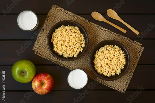 Honey flavored breakfast cereal in two rustic bowls with glasses of milk, apples and wooden spoons on the side, photographed overhead on dark wood with natural light photo