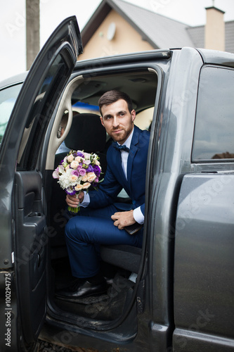 Happy handsome groom with a beard, getting out of wedding car holding a bouquet