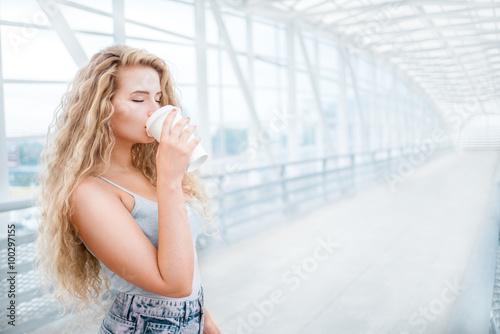 Coffee on the bridge / Beautiful young woman with long curly hair, holding a take away coffee cup and standing on the bridge against urban background.