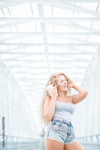 Morning rhythm / Beautiful young woman wearing music headphones, standing on the bridge with a take away coffee cup and posing against urban background.
