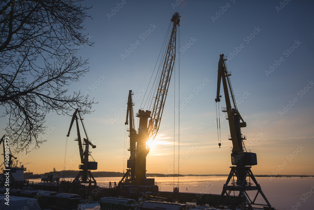 Silhouettes of cranes and industrial cargo ships in port at sunset or dusk, sea port with cranes and docks, in a sunny winter day, back light silhouettes
