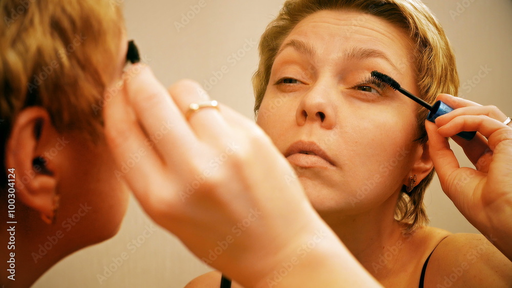 Pretty blond woman applying mascara make-up on her eyelashes in front of mirror and looks at herself. 4K UHD video footage. Pretty blond woman applying mascara make-up on her eyelashes in front of mirror and looks at herself. 4K UHD video footage.