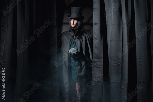 Witch fashion woman wearing black cape and hat. Holding glass sp