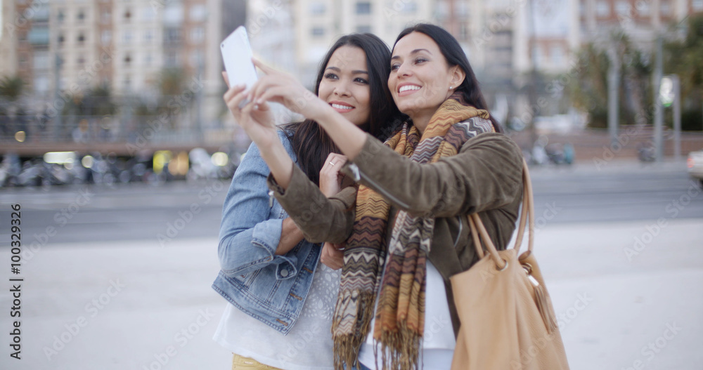 Two gorgeous women posing together for a selfie on a mobile phone as they stand outdoors in an urban square or park
