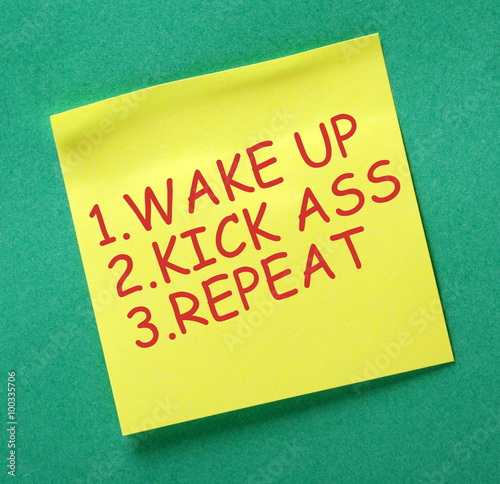 Yellow sticky note with the words Wake Up, Kick Ass and Repeat in red text as a motivational reminder