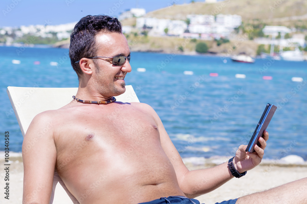 young man laying on a sunbed on the beach relaxing and reading with a tablet