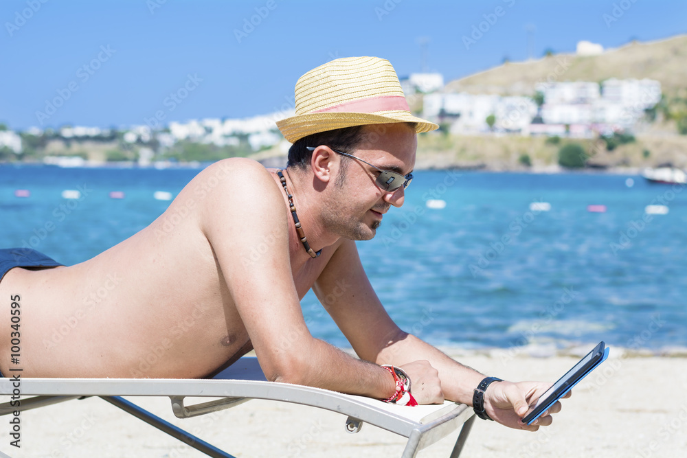 young man laying on a sunbed on the beach relaxing and reading with a tablet