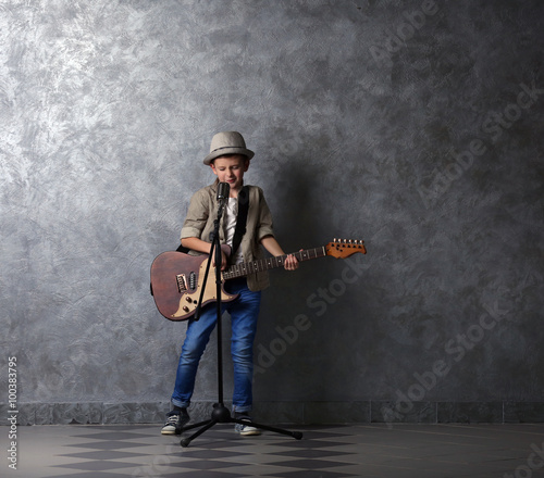 Little boy playing guitar and singing with microphone on a grey wall background