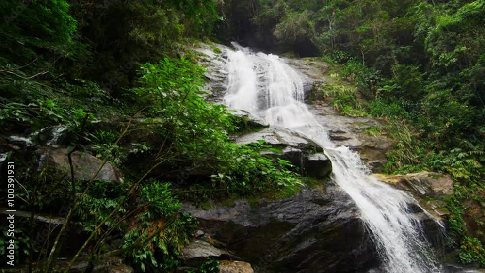Slow motion shot of a jungle waterfall cascading down a dark rocky outcropping into a green pool. Slow motion shot of a jungle waterfall cascading down a dark rocky outcropping into a green pool.