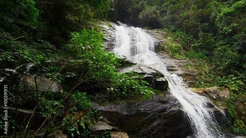 Slow motion shot of a jungle waterfall cascading down a dark rocky outcropping into a green pool. photo