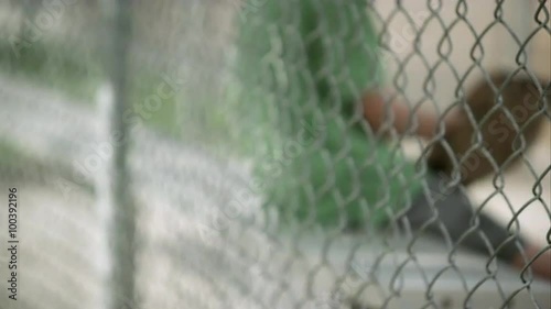 Slow motion rack focus of boy sitting in dugout behind chain link fence. photo