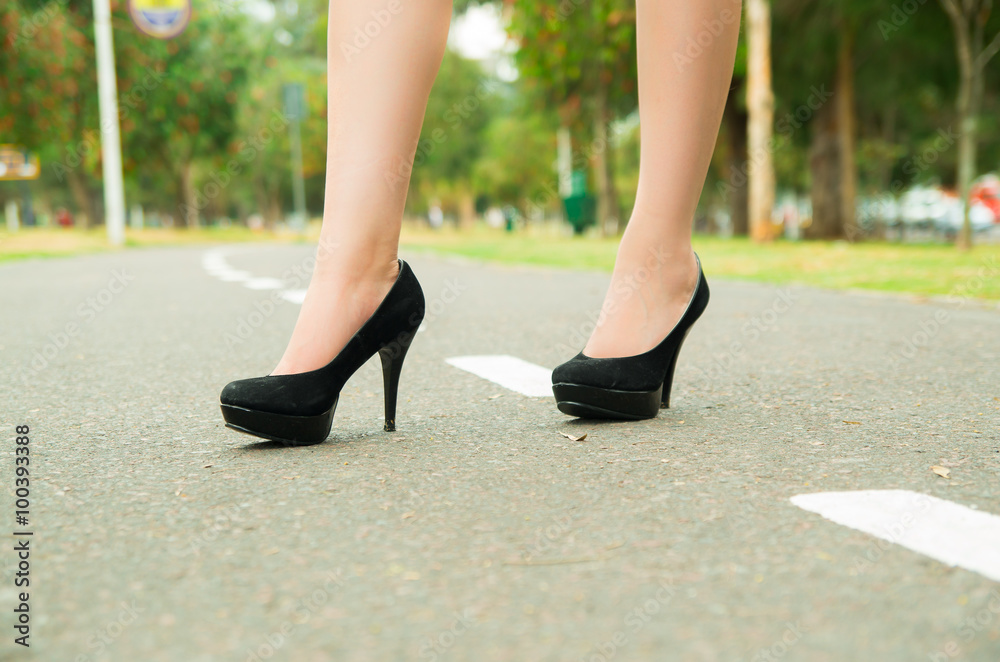 Elegant feet wearing black high heels walking on pavement, green park environment background