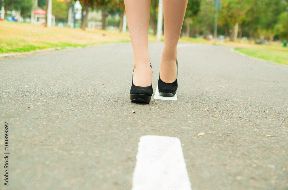 Elegant feet wearing black high heels walking on pavement, green park environment background