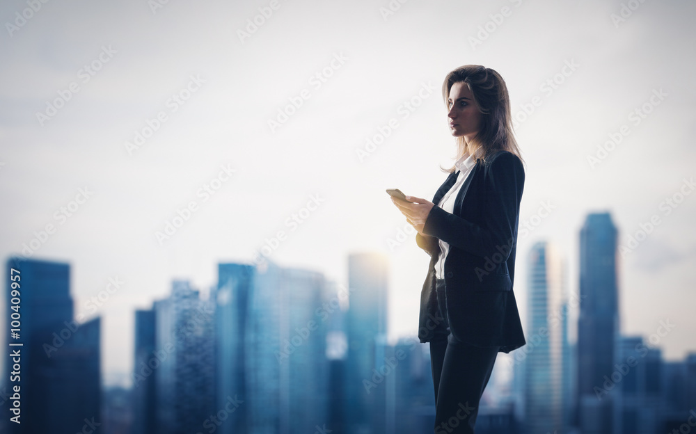 Young woman wearing modern suit holding her smartphone in a hands. City on the background Young woman wearing modern suit holding her smartphone in a hands. City on the background
